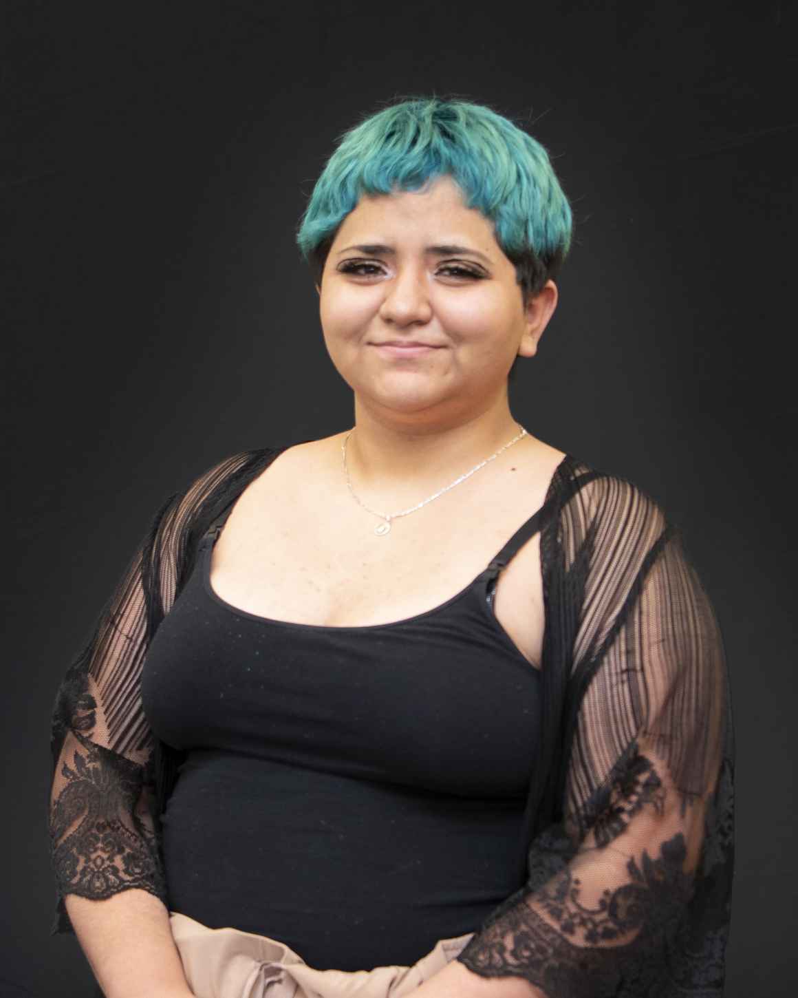A photo of Cristina from the waist up against a black background. Cristina has short green hair and is wearing a black tank top with a black shawl. 