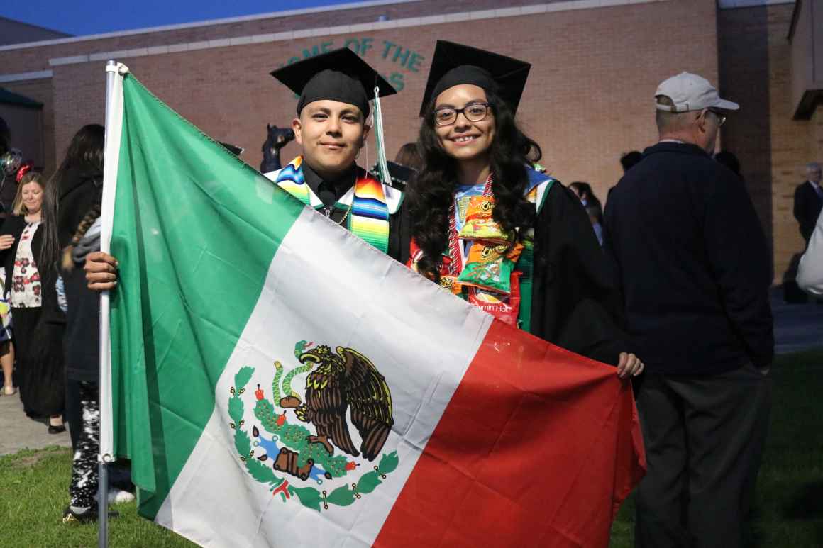 A picture of Damaris and another person wearing graduation caps and gowns. They are holding a large Mexican flag in front of them.