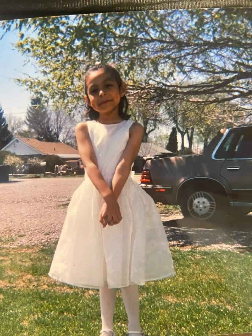 A picture of Damaris when she was young girl. She is standing outside wearing a white dress with her hands crossed in front of her. 