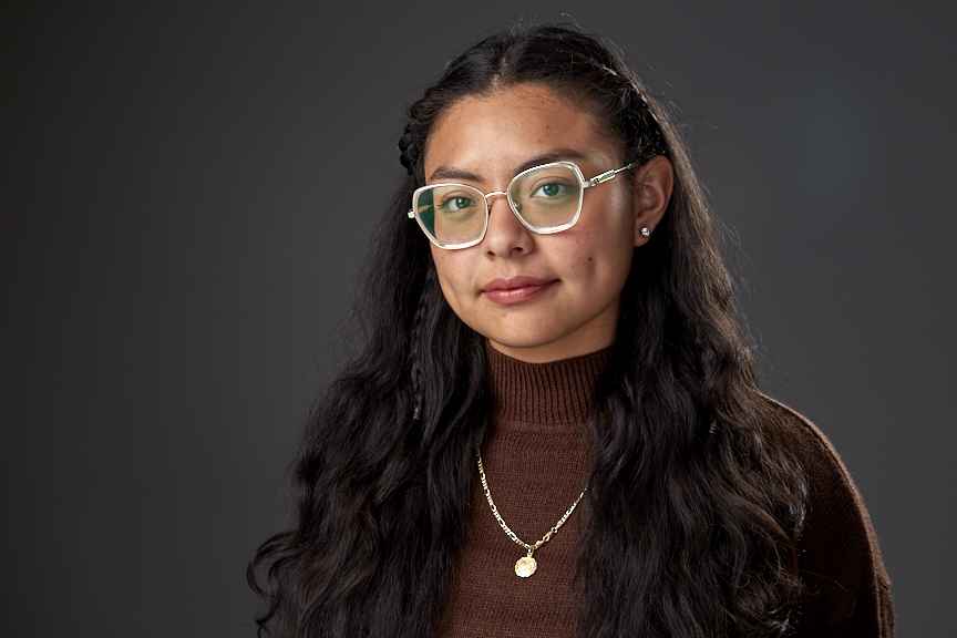 a pictuure of Damaris against a grey background. Damaris is wearing a brown sweater and has silver rectangle glasses.