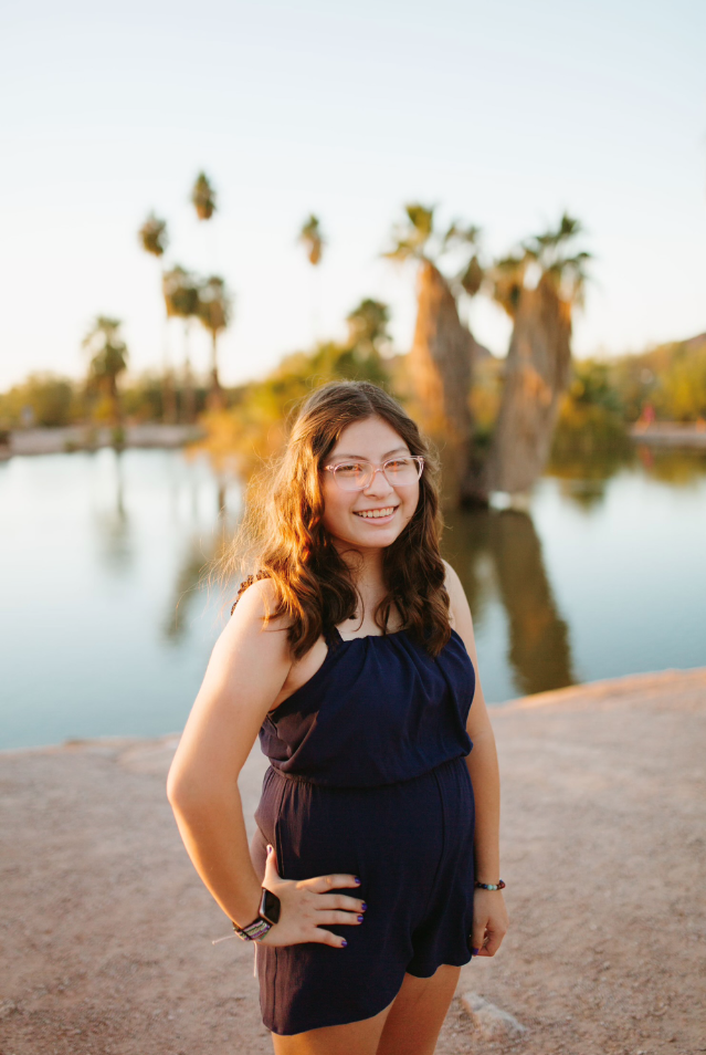 A picture of Jennifer standing in front of a pond with palm trees around it. She wears glasses and has on a navy blue romper. She is standing confidently, looking at the camera with her right hand on her right hip.