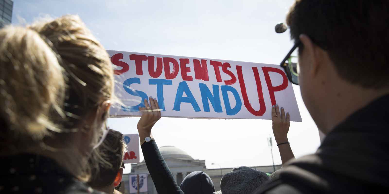a person is holding up a white banner that reads "students stand up" in read and blue letters. In the foreground there are two people with their backs to us to either side of the sign. 