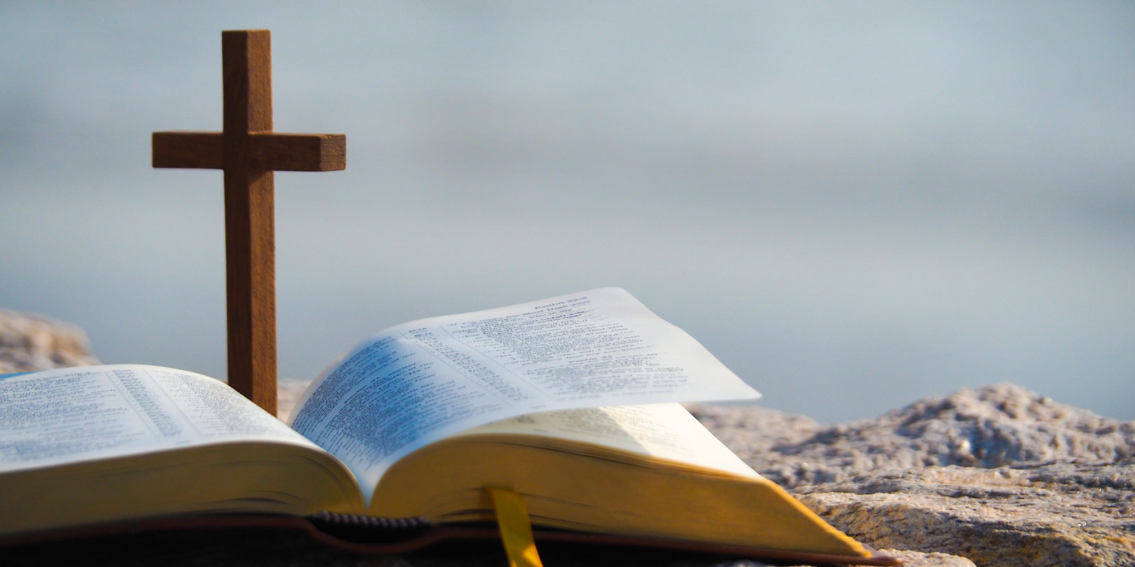 A wooden cross stands up on top of a Bible, both sitting on a rock with a blurred background