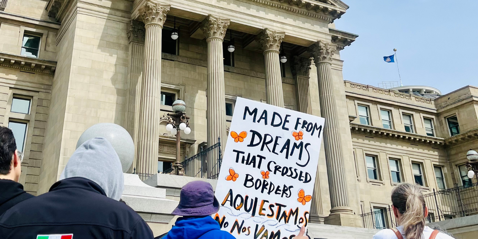 A person holds a sign outside the Idaho Capitol building that reads "made from dreams that crossed borders. aquiestamos no nos vamos"