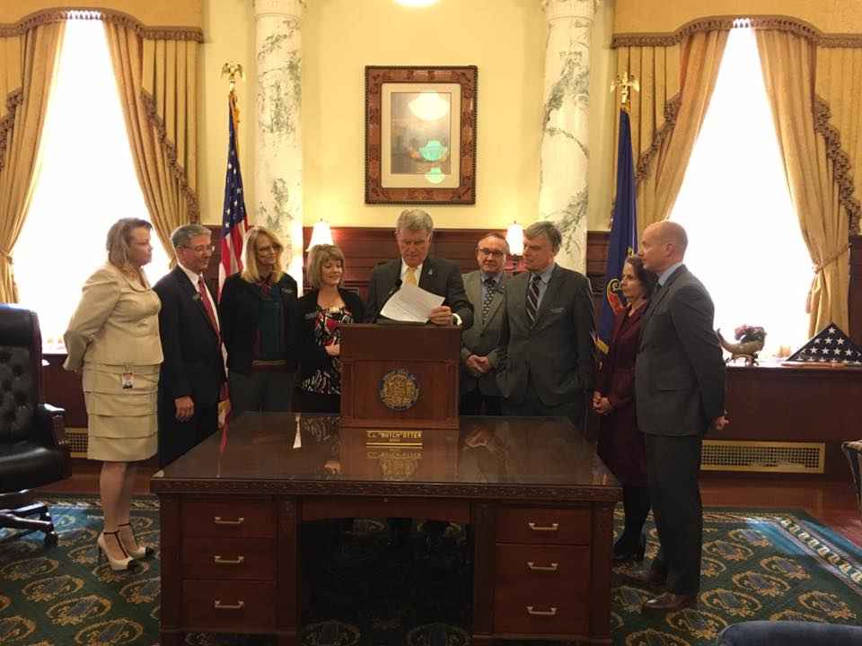 Gov. Otter standing at a podium signing a bill into law in the Governor's office with people standing around him