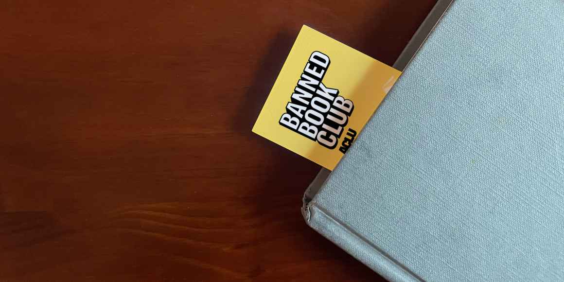 The top left corner of a blue book sits on a brown surface. A Yellow bookmark stuck our of the top of the book with the words "Banned Books Club ACLU" in white and black letters. 