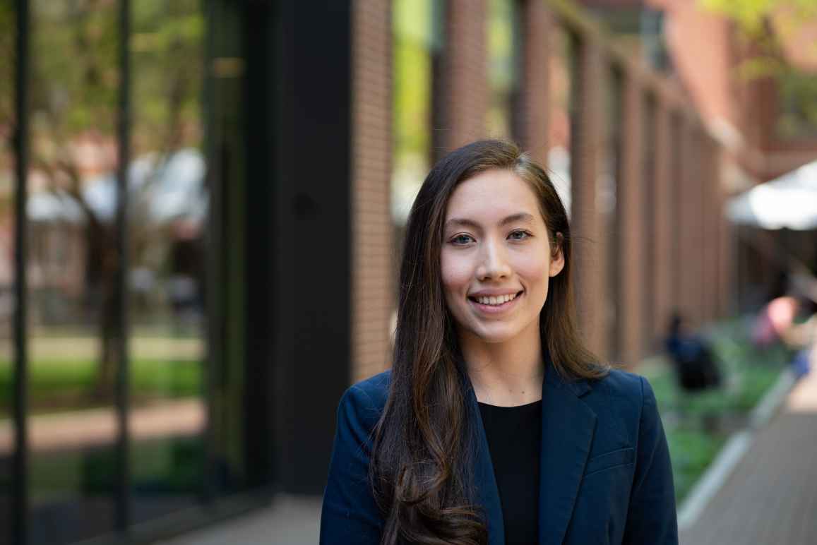 Erica is just right of center and standing outside of a buiding with windows. The background is blurry. She wears a dark blue blazer with a black shirt.