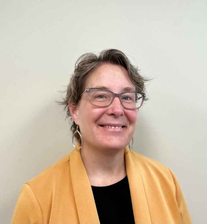 A phot of Julianne wearing grey framed glasses, round earrings in a yellow blazer with a black shirt. Julianne is smiling. 