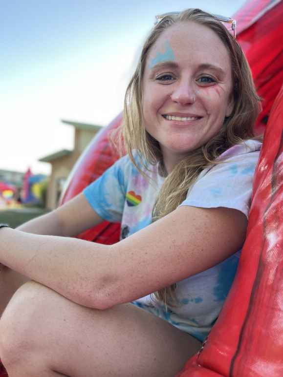 A picture of Jenna sitting down with their arms resting on their knees. Jenna is smiling and wearing a tie-dye t-shirt. Jenna has colorful face paint above their left eye and below their right cheek.