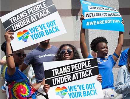 A group of young black people hold up signs in the sunshine that say "trans people are under attack, we've got your back"