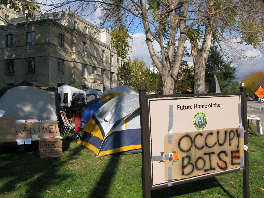a sign with a cardboard sign taped on top with spray painted "occupy boise" on it with tents in the background