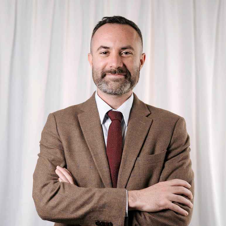 A photo of Paul against a white wavy backdrop. He has short dark hair and had a beard. He has his arms crossed in front of him and is wearing a brown suit jacket with a white shirt and maroon tie. He is smiling.