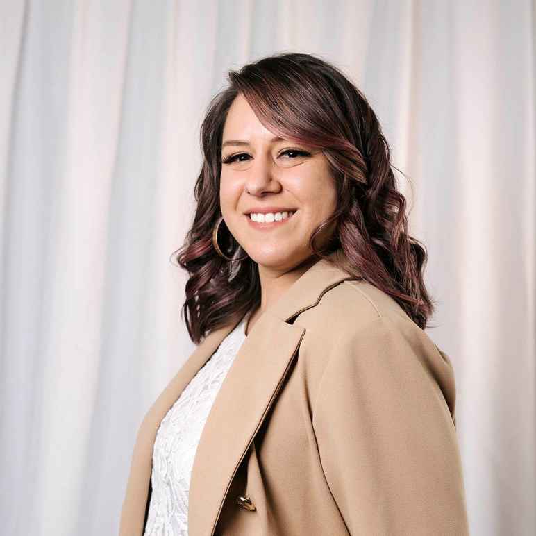 A photo of Rebecca. She has long dark curly hair and is wearing hoop earrings, a beige sport coat and a white shirt. She is smiling and is set against a white wavy backdrop. 