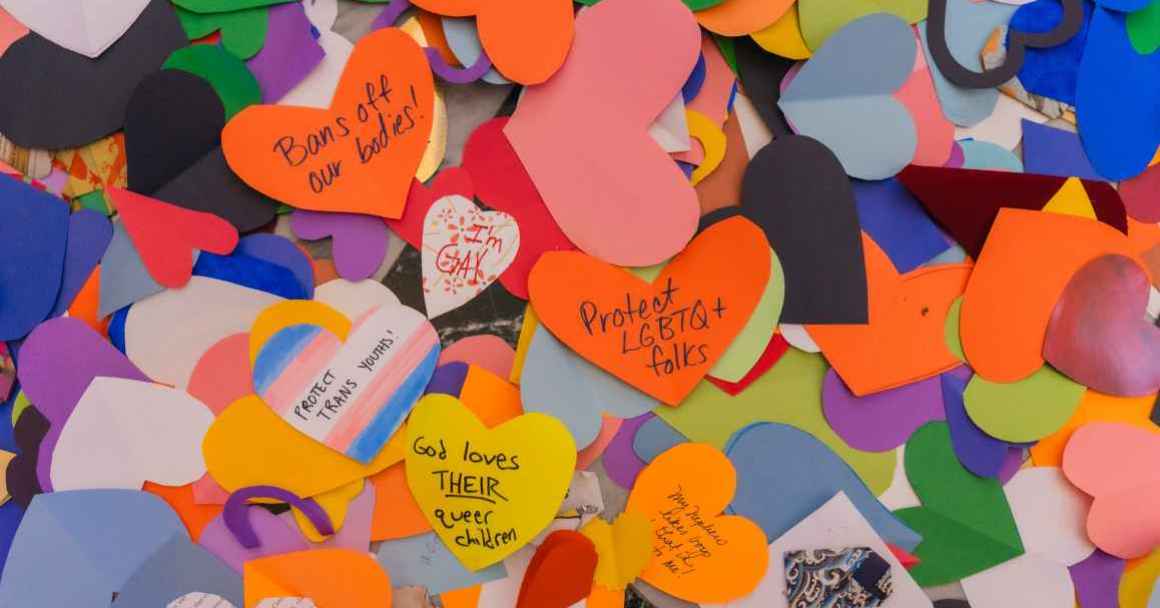 Collection of paper hearts in a variety of colors sharing uplifting messages for the LGBTQIA+ community.