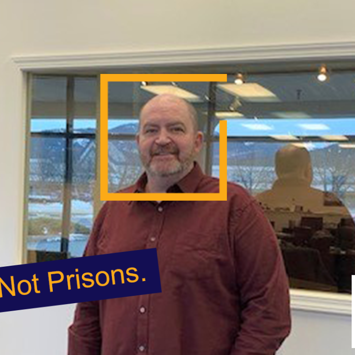 John is a middle-aged balding white man looking at the camera and smiling from in front of a business's wall and window into an office.